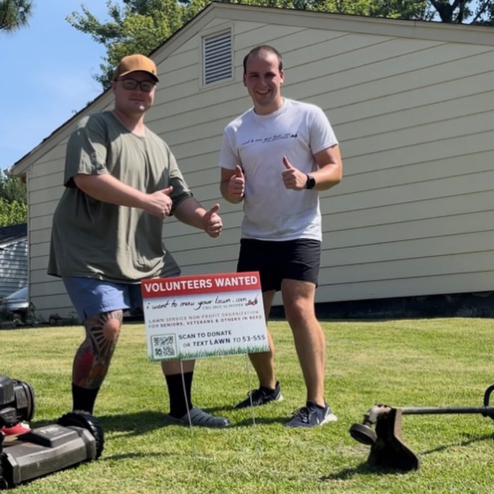 Active-duty Navy volunteer Max giving a thumbs up next to a Volunteers Wanted yard sign during an IWTMYL community service visit