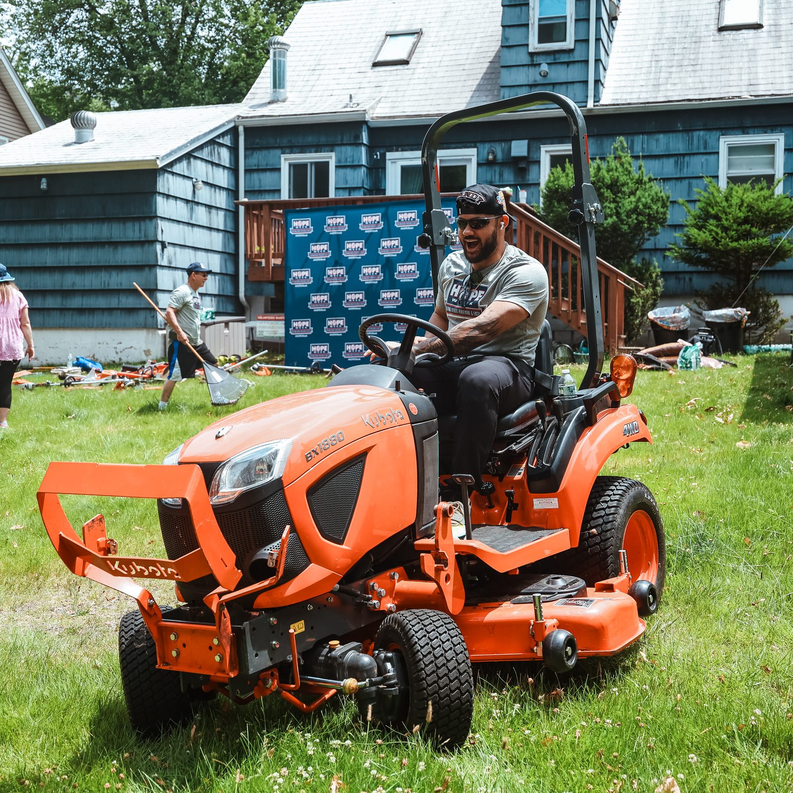 Jasson Dominguez drives an orange riding mower across the lawn in front of a blue HOPE Week backdrop.