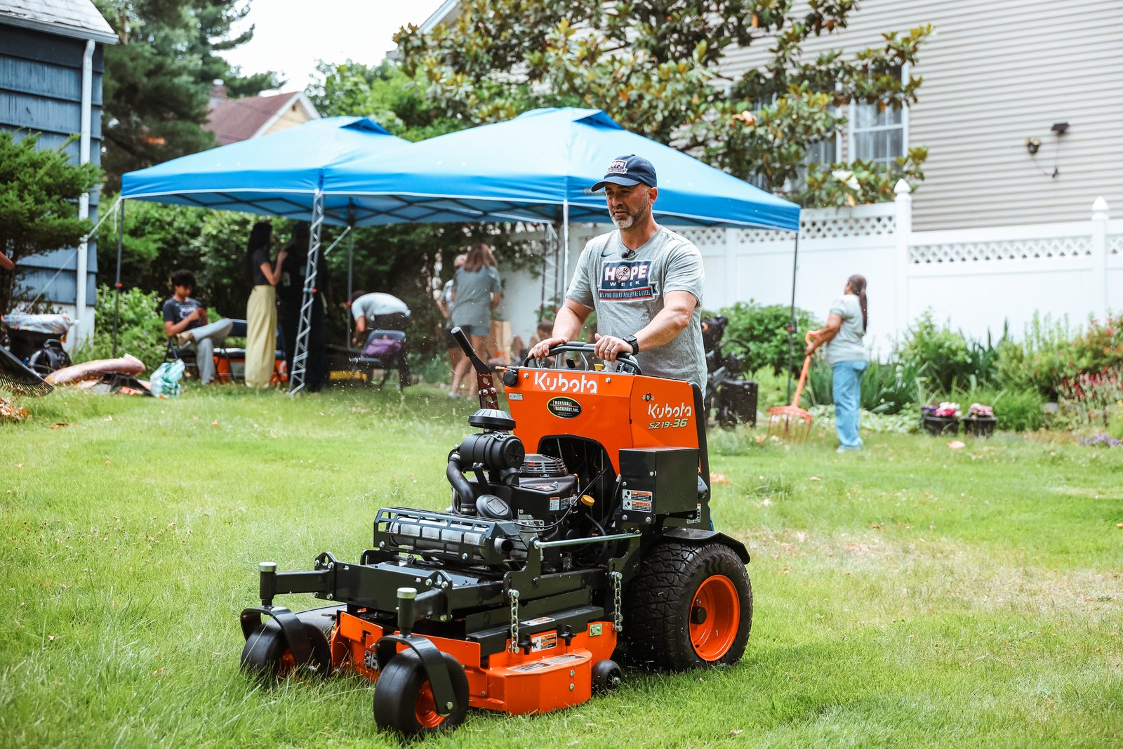 Brian Schwartz drives an orange stand-on mower across a freshly cut back lawn, with a blue event canopy and other volunteers behind him.