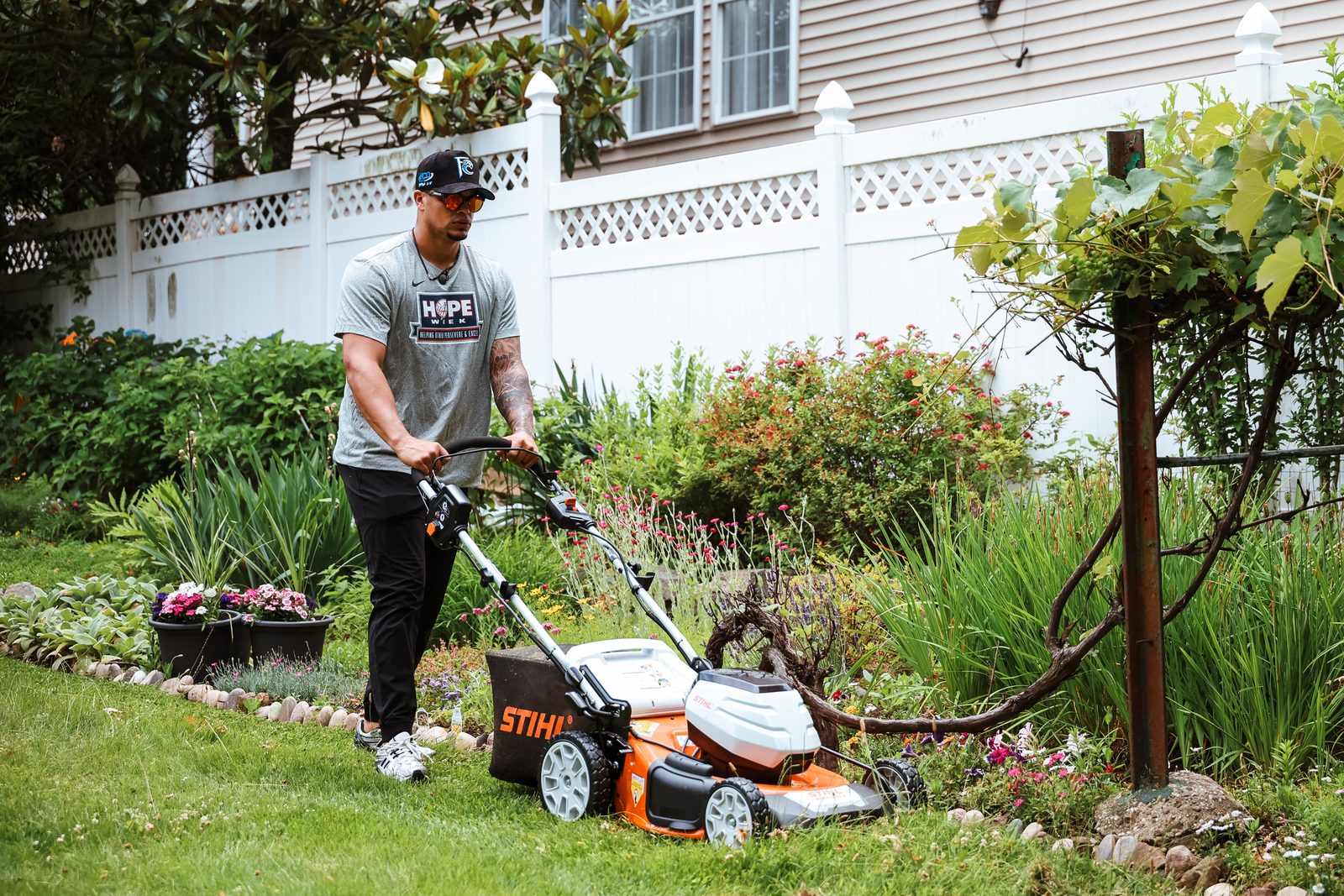 Fernando Cruz pushes a walk-behind mower along the edge of a planted garden bed beside a white fence.