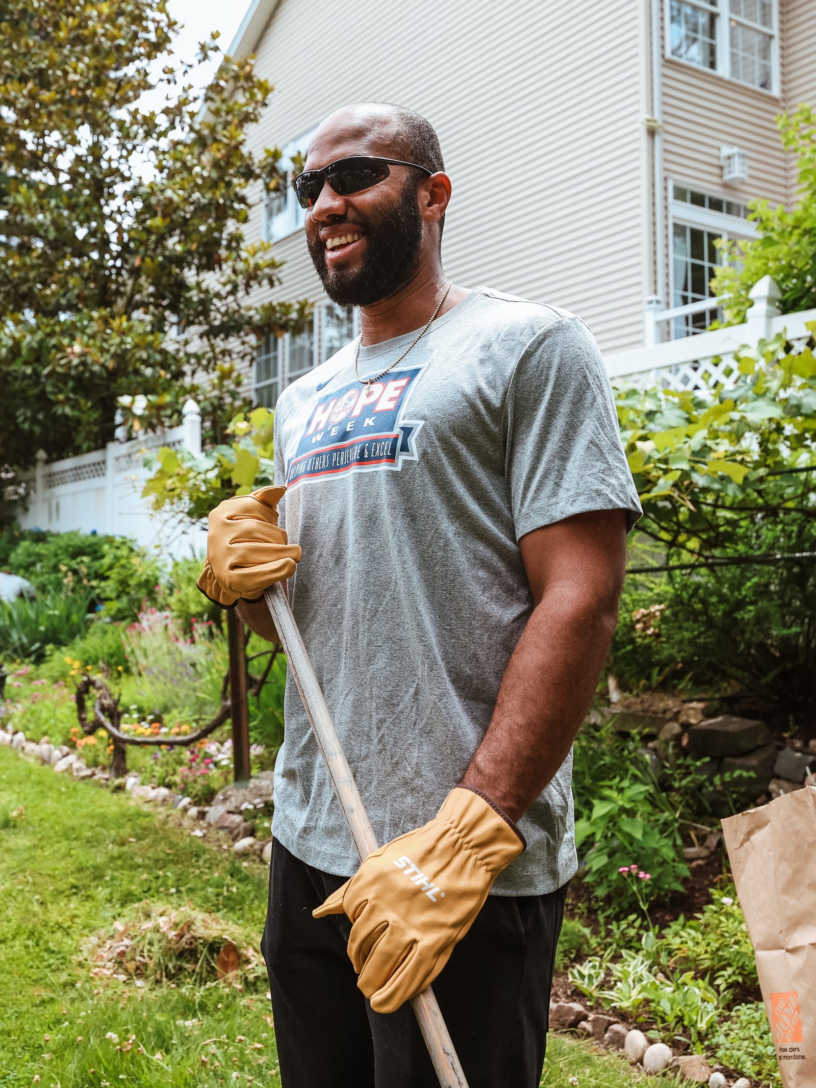Amed Rosario, in work gloves and a HOPE Week T-shirt, smiles while leaning on a rake beside a garden bed.