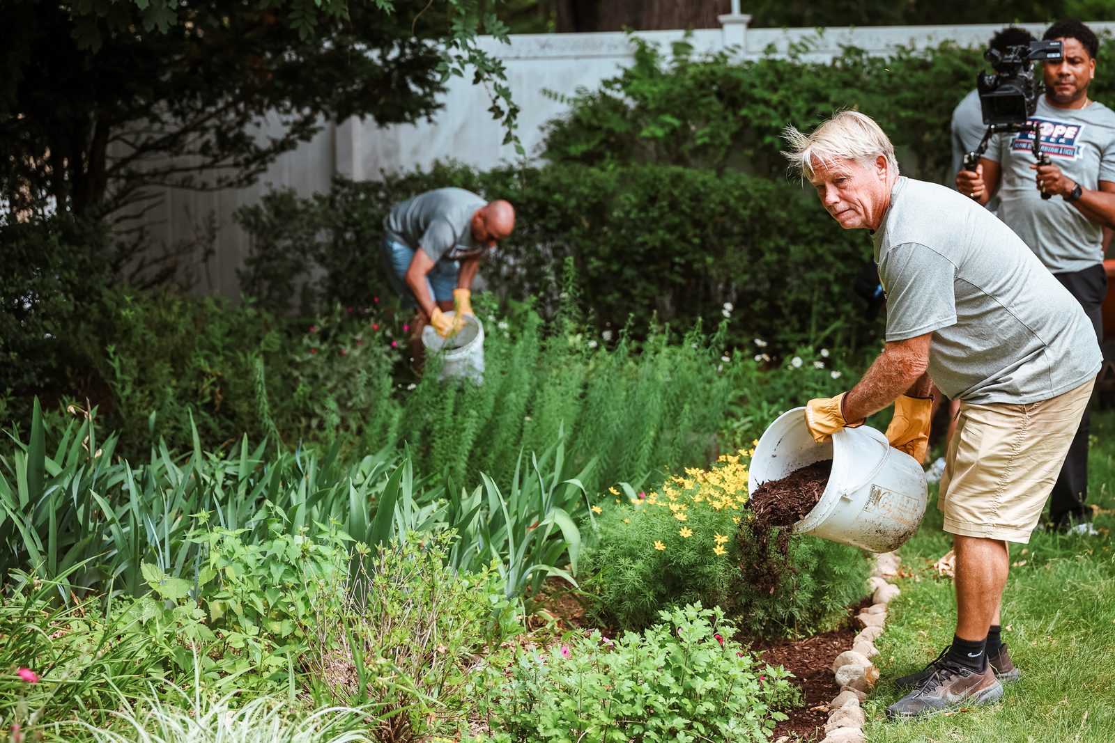 Danny Cunningham tips a bucket of mulch onto a garden bed while a camera operator films nearby.