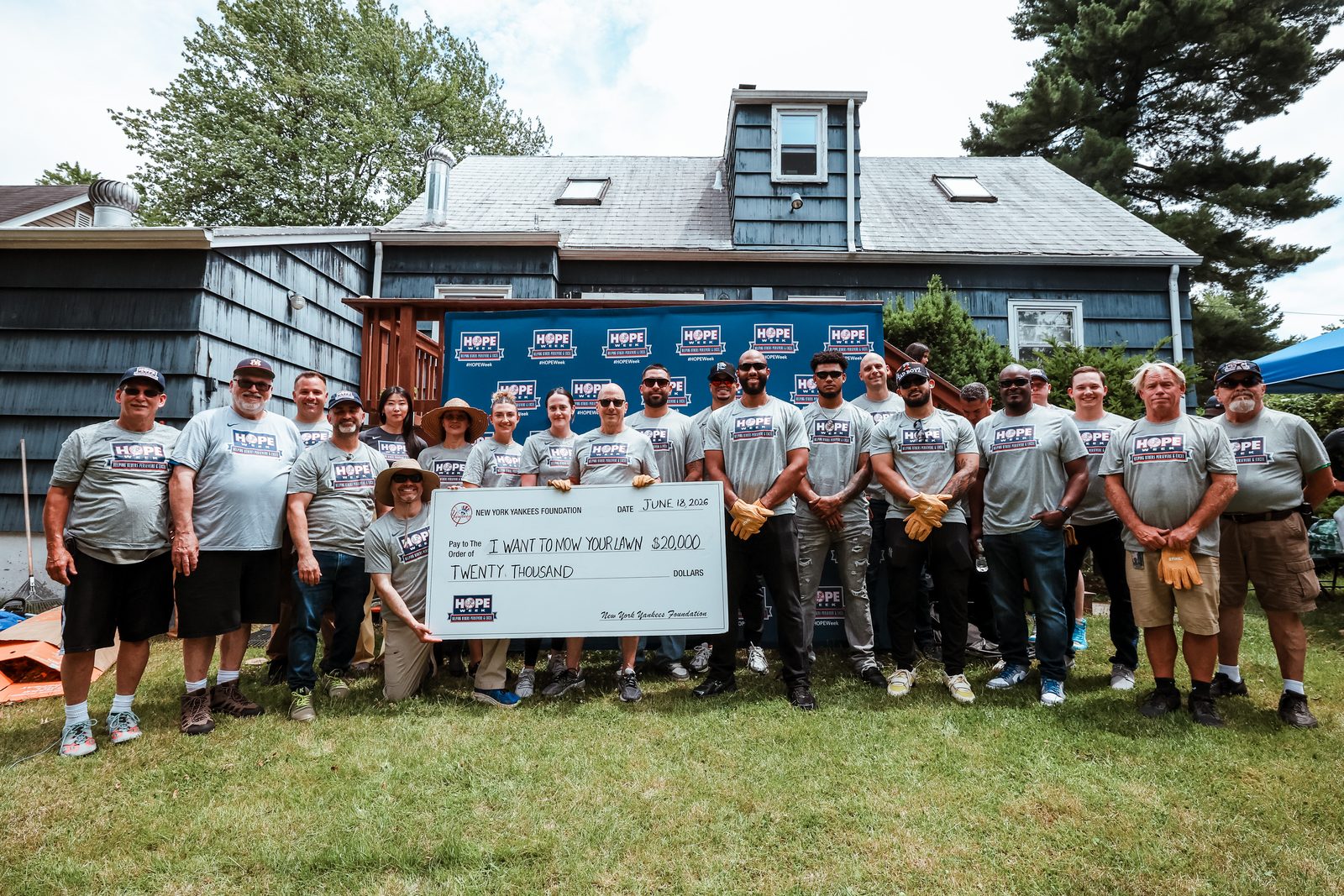Yankees staff and I Want To Mow Your Lawn volunteers in HOPE Week T-shirts stand together outside a blue-shingled house for the HOPE Week honoree presentation, holding an oversized ceremonial check.