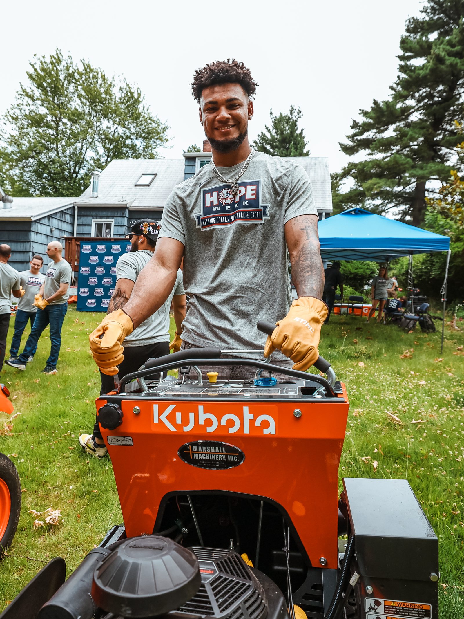 Camilo Doval, wearing work gloves, stands behind an orange mower and smiles toward the camera.