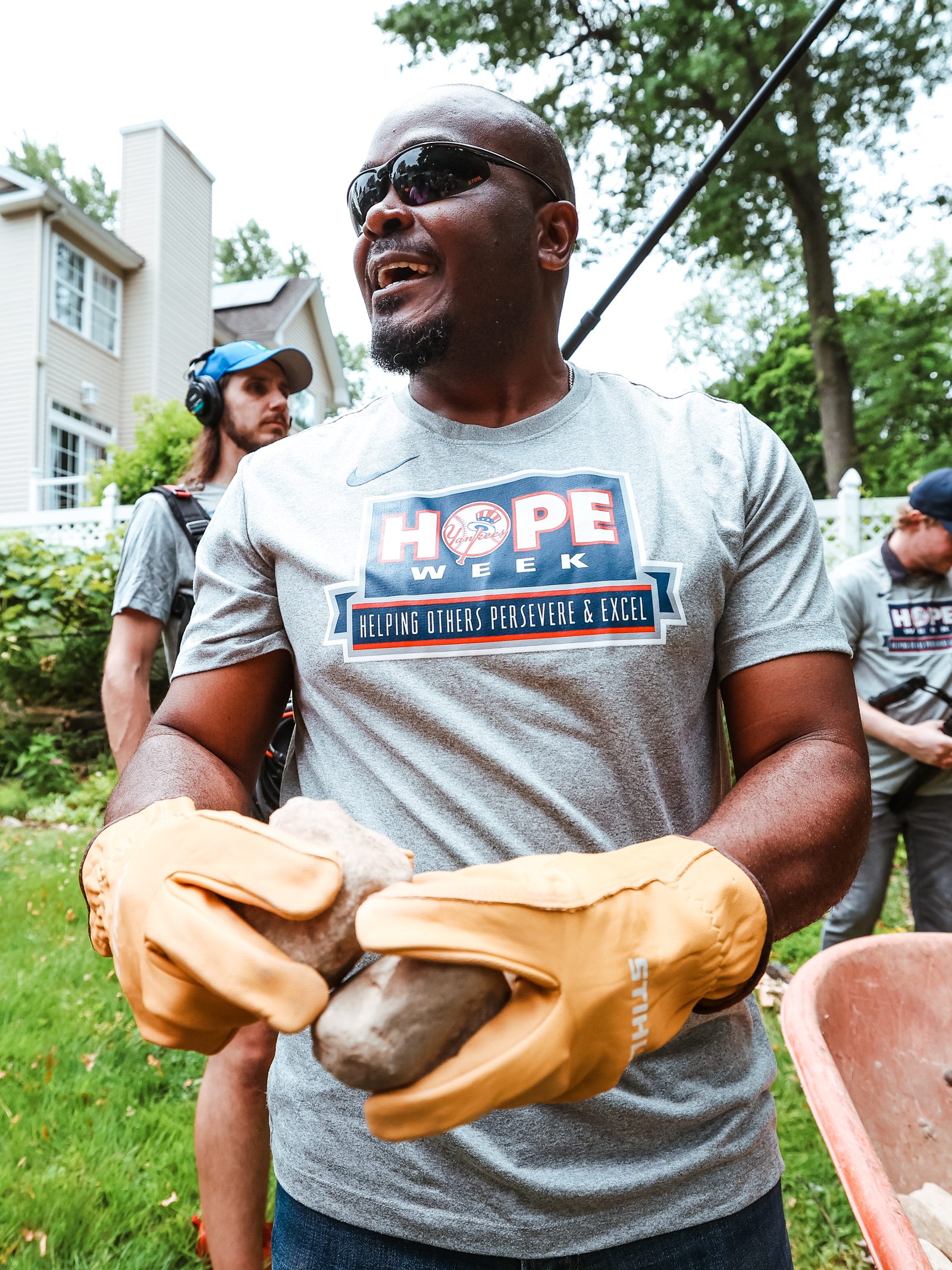 James Rowson carries a large garden stone in gloved hands, wearing a HOPE Week shirt reading Helping Others Persevere and Excel.