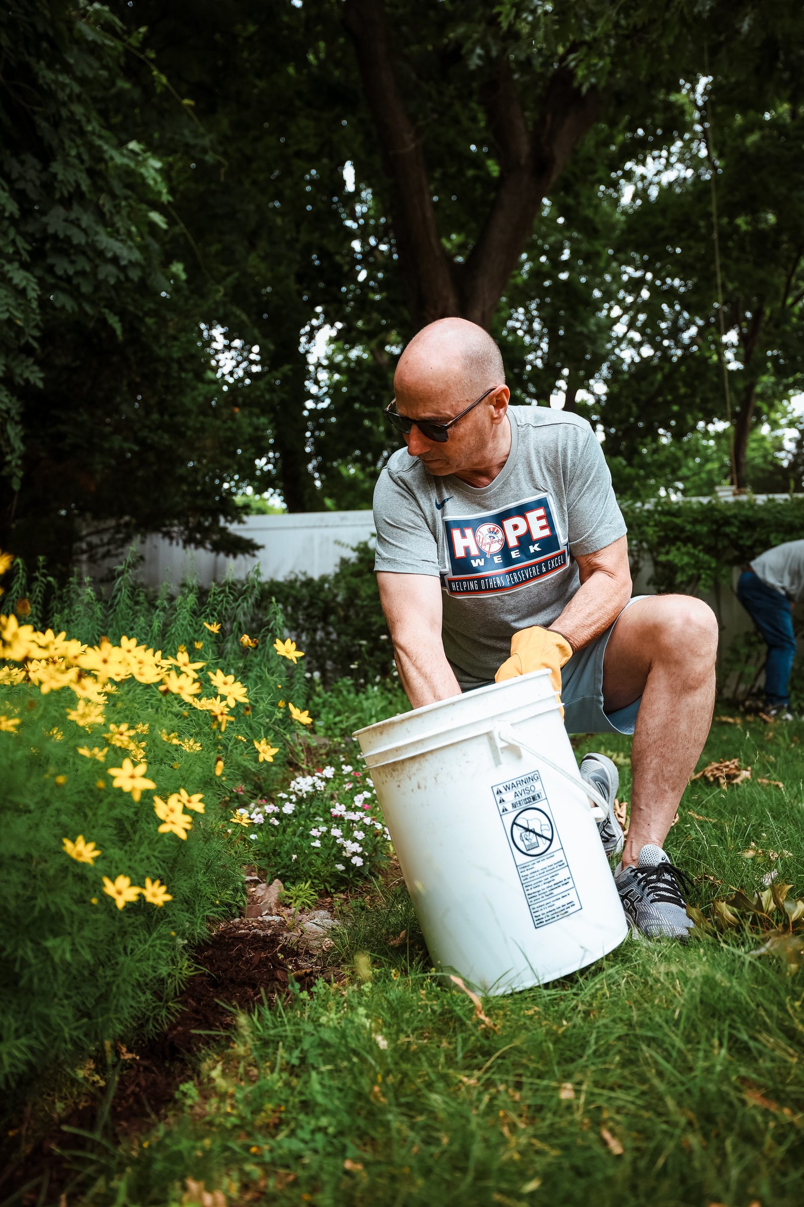 Brian Cashman crouches to empty a bucket of mulch into a flower bed edged with yellow blooms.