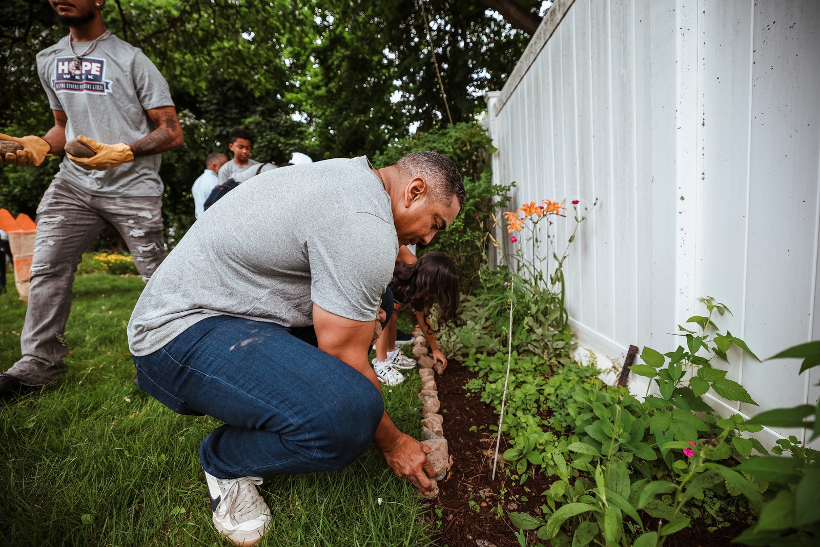 Luis Rojas crouches to set plants and stones into a freshly mulched bed along a white fence.